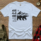 White t-shirt with black bear, mountains, forest, and "B.C.D." text.