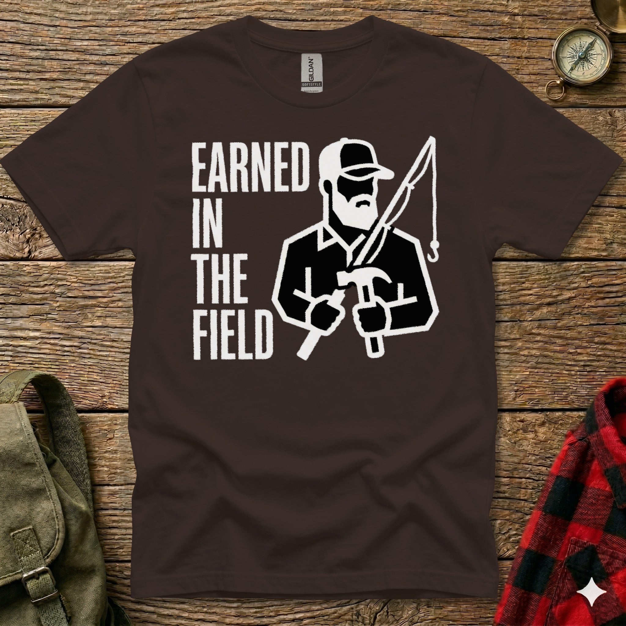 Brown t-shirt with "EARNED IN THE FIELD" and a graphic of a man fishing and hammering.